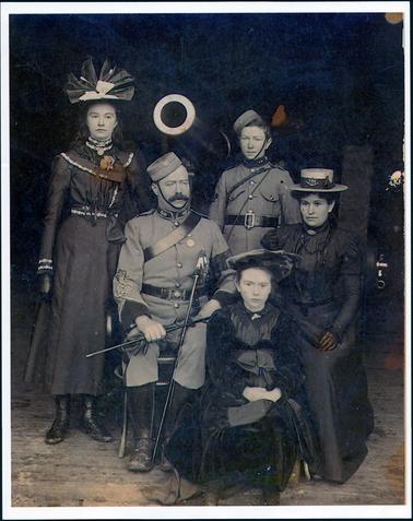 This image shows a group of people posing for a portrait.  Two young people are standing at the rear, a man and a woman are sitting and a young girl is seated in front of them.  The man is dressed in full military uniform.  The woman is wearing a dark full-length dress.  It has a belt at the waist, a high neckline, with a brooch, a half collar and puffed long sleeves.  There is lace on the collar and on part of the bodice.  She is wearing a straw hat with a wide band and a brooch attached at the front.  The young woman standing is wearing a three-quarter length, long sleeved dress with a large collar and a high neckline with a bar brooch attached.  There is lace around the collar, neckline, wrist and waist which has a sash tied in a large bow.  Her hair is long and she is wearing a matching high hat with a large white bow at the front with two hat pins.  She is wearing black boots.  The other young person, standing, is dressed in a military uniform.  The young girl sitting is wearing a dark coloured, long sleeved dress with a lace collar and a brooch attached.  She has a very wide sash tied with a big bow at her waist.  She is wearing a hat and boots.  A large gun barrel can be seen in the background.  There are old, bare floorboards on the ground.  