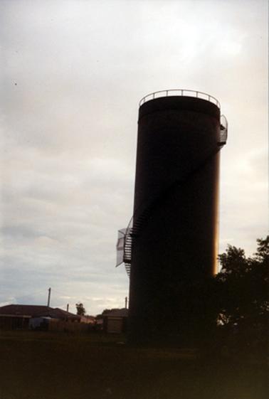 This photograph shows a large water tower at the centre of the image with trees to the right and houses to the left.  The tower has steps going from the bottom left of the tower, across the middle to the top right.  A railing is around the top of the tower.