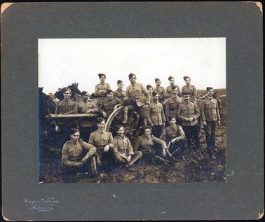 This image shows a group of soldiers posing for a photograph around a cannon.  There are nineteen men.  Five are sitting on the ground in front of the cannon, one is sitting on the wheel of the cannon and the remainder of the men are standing.  Horses can be seen on the left-hand side of the image and more men and horses can be seen on the far right-hand side of the image.  All the soldiers are dressed in the same uniform except the man, second from the right, who appears to be an officer.  The photographers details are embossed at the bottom left-hand side of the mounting board.  