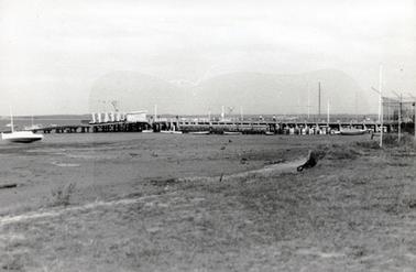 This image shows a wooden two-masted ketch with a jetty in the foreground.  Three men and a dog can be seen on board.  Fishing nets and boxes cover the floor of the boat.  A strip of land can be seen in the background.  
