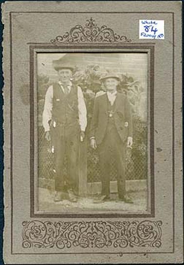This image shows two men standing on a path posing for a photo.  Both men are very well dressed in three-piece suits, although the man on the left has removed his coat.  Both men are wearing hats.  They are standing in front of a wire fence with tall shrubs and a brick wall behind them.  There is a white sticker on the front top right-hand side of the mounting which reads: “White 84 Family No”. 