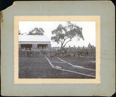This image shows a large group of men in uniform posing for a photograph.  They are in a grassed paddock in front of a large tree.  A shed with two large open doors is behind them on the left-hand side of the image.  Large trees are behind the shed.  There is a large gun on a gun-carriage in the centre of the image with about ten men pulling a rope attached to a wheel of the carriage.  Other men are standing around.  Six men are standing to attention, in a line, on the right-hand side of the image and a greyhound dog is behind them.  A soldier on horseback is behind them.  Part of another gun-carriage can be seen behind him.  Planks that the gun-carriage run on, are seen on the ground.  