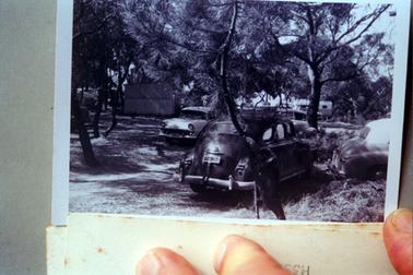 This image shows four motor cars parked underneath trees.  A caravan and other motor vehicles can be seen in the background.  A small building is underneath the trees on the left-hand side of the cars.  The number plate on the car in the foreground is: FB 394. 