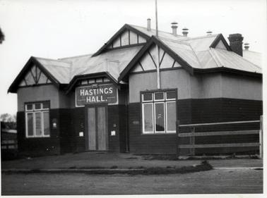 This image is a photograph of a hall which has the words ‘Hastings Hall’ printed in large white letters above the front door.  The bottom half of the building is constructed of timber.  The roof-line has several triangular shapes with detailed sections.  There is an unusual shape in the roof-line over the door.  Three sets of windows are on either side of the door.  There is a brick chimney on the roof along with several  small pipe outlets.  A post and rail fence is on either side of the building.  A footpath and a road are in the foreground.  A flag pole is above the set of windows on the right-hand side of the photograph.  