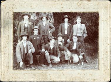 This image is of a group of nine men in front of a large tree trunk.  Four men are standing at the back and five are kneeling/crouching at the front. The last man on the right-hand side of the photo has one arm around a dog and he is holding a stick in his other hand.  All men are dressed in three-piece suits.  Seven of the men are wearing hats and the other two men are wearing caps.  Five of the men have flowers in their lapels.  Small trees are behind the large tree trunk.  