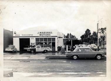 This image shows a square building with a high open door on the left-hand side.  A large glass shop-front window and a small doorway are on the right-hand side of the building. Shelving can be seen behind the glass.  The back of a car can be seen inside the building.  Three petrol bowsers are at the front of the building on the right-hand side.  Several cars are parked at the front and side of the building.  A street is in the foreground .  Part of a street can be seen on the far right-hand side with other buildings and tall trees.  Signage across the front of the building reads as follows: “Esso Servicenter J. Marshall Motors Licensed Vehicle Tester”.  The utility parked at the front of the building has ‘National Tyre Service’ printed on the side.  