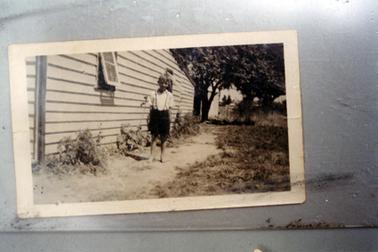This image shows a small boy standing on a pathway beside the wall of a weatherboard building, posing for a photo.  He is wearing a white short sleeved shirt and black shorts with braces.  He is barefoot.  The building has two open windows along the wall.  Small shrubs are along the wall and tall trees are in the background on the right-hand side.  