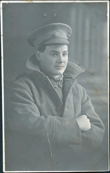 This image shows a soldier in uniform, including cap and coat, posing for a photograph, from the waist up.  The collar of his coat is standing up, his arms are folded and he has a slight smile on his face.  He is standing slightly side-on.  