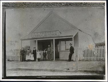 This image shows a shop with a group of people standing outside posing for the camera.  On the left of the photograph is a man with a bicycle.  Next to him a woman, nursing a baby and a small child is standing next to her.  Next is a man wearing a long black coat, a small girl, a small boy and a man wearing a striped butcher’s apron.  A man is leaning against the post on the far right of the photograph.  The sign on the shop reads: “F.G. Perrott Family Butcher”.  The shop is of timber construction with a large glass window to the left of the open front door and a large opening in the wall to the right of the door.  It has a front verandah with four wooden posts.  A picket fence is on either side of the shop.  There is a footpath, a gutter and part of the street in the foreground.   