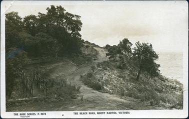 This postcard shows a winding dirt road climbing a hill.  Native trees and shrubs are on either side.  A bank is on the left-hand side and the land slops down to the water on the right-hand side.  A wooden foot bridge is in the foreground on the left.  Printed across the bottom of the postcard is: “The Rose Series, P. 2074 Copyright The Beach Road, Mount Martha, Victoria”.  