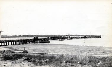 This image shows a jetty at low tide.  There are several fishing boats tied up alongside.  Tuffs of grass can be seen in the foreground and part of a shed can be seen on the far left-hand side of the image.  Several tall poles line the jetty.  Land and trees are in the background. Two people can be seen standing on the jetty.  