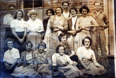 This image shows a group of fifteen people posing for a photo.  There are three men and six women standing and six women sitting on the ground in front of them.  They are all smiling for the camera.  The two men at each end of the back row have their shirt sleeves rolled up.  The other man is wearing braces.  The women are wearing either skirts and tops or print dresses.  Some are wearing cardigans.  Part of a brick wall can be seen behind the group.  A window is on the left-hand side of the wall.  