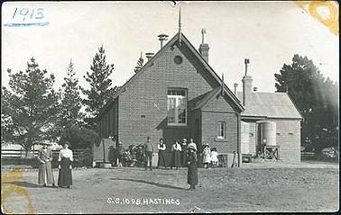 This image depicts Hastings State School no. 1098 in 1913. The side of the brick building is shown, with a brick annexe to the right side and a corrugated iron roof. There are 8 adults and a small group of children  grouped in front. There are 2 corrugated iron water tanks attached to the side annexe of the building. At the bottom of the image is written in white “S.S. 1098 Hastings”