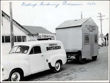 This image shows a white Holden panel van, presumably in the main street, with the words “Perrott & Co. Butchers” on the side of the van. There is one man, the driver, in the van. The van is towing a trailer carrying a small mock house with the words “F.G. Perrott family butcher est 1883” written on the side. Behind the van is a small timber shop with the words “New & used furniture” on the side of the verandah. Another small timber building can be partially seen behind the trailer, with a man and a woman standing in front.