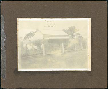 This image shows a weatherboard cottage with a four post verandah at the front.  Two windows are on either side of the front door and one window can be seen on the side of the cottage. A brick chimney is on the right-hand side of the roof.  A post and wire fence is across the front with a small gate at the right end.  Shrubs are in the garden and tall trees can be seen behind the cottage.  Bare ground is in the foreground.  Printed at the top of the photograph is: “Rose Villa Hastings”.