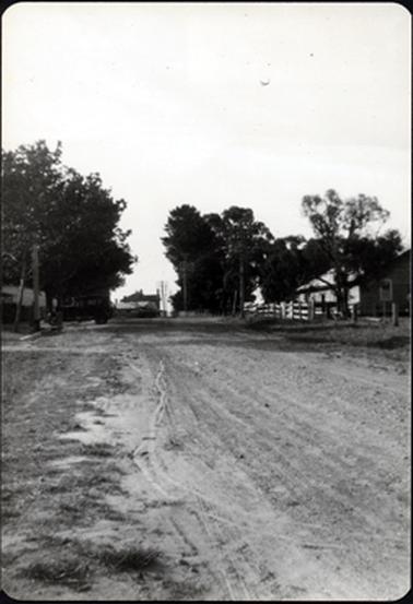The bottom half of this image shows a gravel road with grass on each side.  A building can be seen behind a post and rail fence on the right-hand side of the image.  Buildings and telegraph poles can be seen in the distance. Part of a fence and building can be seen on the left-hand side of the image with a parked car across the footpath.  Tall trees are on either side of the street.