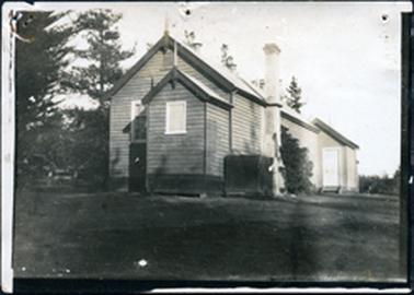 This is an image of a school.  It is a weatherboard building with an attached front entrance.  There is a large chimney on the right-hand side of the photo.  There is a small square tank near the chimney.  A small building with a door can be seen at the back of the school. There are large pine trees on the left-hand side of the photograph and trees or shrubs in the distance on the right-hand side of the photograph.