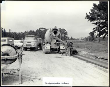This image shows works in progress on a street.  In the centre of the image a cement mixer is pouring cement to form the street gutter.  Three men are seen working the equipment.  An Austin truck, number plate NVD483 is nearby.  The rear of two vehicles can be seen on the far left-hand side of the photograph.  A hand cement mixer is in the foreground.  A grassed embankment is on the right-hand side of the photograph and trees are in the background.  Typed on a piece of white paper and glued to the bottom of the photograph is: “Street construction continues”.