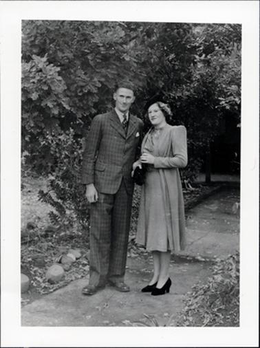 This image shows a man and woman standing on a concrete path is a garden, posing for a photo. They are both very well dressed.  The man is wearing a checked three piece suit, white shirt and tie.  The woman is wearing a plain coloured knee-length dress with a matching coat.  She is wearing a small black hat, black clutch bag and black shoes.  A rough garden is on either side of the path.  Stones can be seen in the garden on the left.  Bushy trees are behind them.  