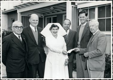 This image shows a group of five men and a nursing sister posing for a photograph outside a building.  All the men are dressed in suits, vests or waistcoats, white shirts and ties.  The nursing sister is in uniform.  Everyone is smiling and the man on the far right-hand side of the image is handing a piece of paper to the sister.  Part of the roof, walls and windows of the building can be seen behind the group. 