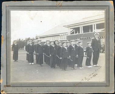 This image depicts a group of young boy cadets (17) on parade with an adult senior officer on right-hand side of photo.  The buildings of the Flinders Naval Depot (now HMAS Cerberus) are in the background.  There are 2/3 Naval personnel seen in the doorway of building watching.  The cadets are holding a rope. Written on the front of the photo “? 25.11.1926” Written on the front of the photo in the bottom right corner: “25.11.1926”. There is something else written but this cannot be read. 