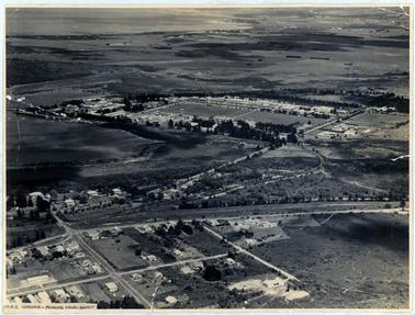 This image shows an aerial view consisting of buildings, roads, paddocks and the shoreline on the left-hand side of the image.  A large section in the centre of the image shows cleared land surrounded by large buildings.  Printed at the bottom left-hand corner is: “H.M.S. Cerberus - Flinders Naval Depot”. 