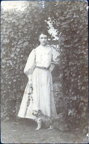 This image shows a beautifully dressed young woman standing under an archway covered in a creeper.  There is a terrier dog standing at her feet.  Her dress is almost to the ground.  It has a beautiful high lace collar, long ‘puff’ sleeves with a wide wrist band.  The top is ‘bloused’ at the waist and the skirt is slightly gathered.  Her hair is parted on the right side of her head.  She is holding a small branch in her right hand.   Wire netting can be seen at the back of the trellis.  