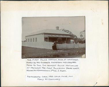 This image shows a small timber cottage with an iron roof and a four posted verandah at the front. The roof of the verandah has a striped effect.  Five windows can be seen along the side of the cottage.  A wooden fence is along the left-hand side of the photograph and a picket fence is across the front with a small gate in the middle.  A large brick chimney can be seen above the roof line.  There are trees on the right-hand side of the photograph where part of a shed can be seen.  There is a woman and child standing outside the fence at the corner of the property and another woman inside of the fence near them. There is a wide grassed footpath and a wide stretch of grassed land beside the cottage.  A sign with the words Hastings Refreshment Rooms can be seen on the wall under the verandah.  Printed on the mounting at the bottom is: “The first Police Station, High St. Hastings. Owned by Mrs Elizabeth Anderson, nee Grayden.  Prior to this, the nearest Police Station was at Dromana.  The First Policeman James Watt, second, Mr O’Shannessy, 3rd Mr J. Egan.  Photograph circa. 1899. Lalla, Anne and Percy McConaghey.” 