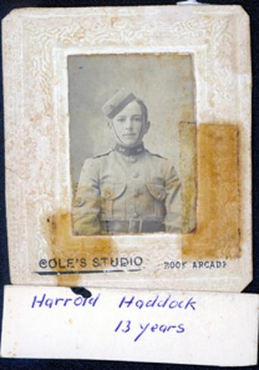 This image shows a portrait of a boy in uniform, taken from the waist up.  His uniform has a small, high, dark, collar at the neck and two large pockets.  He has a wide, dark belt and a pillbox hat with a chin strap.  Printed at the bottom on the mounting is “Cole’s Studio Book Arcade”.  Attached to the mounting with tape is a piece of card that reads: “Harrold Haddock 13 years”.  There are dark marks where sticky tape has been removed.  The mounting has an embossed pattern.  