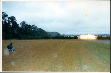 This image shows a man crouching on the grass with his back to the camera, setting off an explosion.  He is wearing a dark jacket and blue jeans. The bottom half of the photograph is green grass.  More grass can be seen in the background behind small trees and the smoke from the explosion.  Tall trees and shrubs can be seen on the left-hand side of the image as well as in the background.  A single tall pole can be seen in the distance in the centre of the image.  