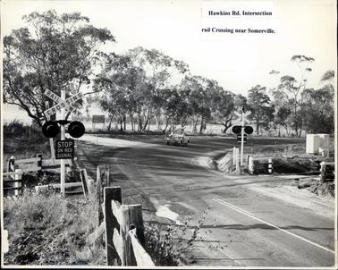 This image shows a railway line crossing a road.  There are railway crossing lights with the sign ‘stop on red signal’ on both sides of the road.  The back of a utility can be seen in the centre of the image driving around a bend in the road.  Several post and rail fences can be seen in the foreground and on both sides of the track.  An advertising sign for “Ron Wilson, Removalist” can be seen on the side of the road between the gum trees which line the left-hand side of the road.  White lines can be seen on the road in the foreground.  Typed in black on white paper and glued to the front of the image is: “Hawkins Rd. Intersection rail Crossing near Somerville.”