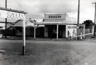 This image shows a street scene with a bakery in the centre of the image.  The bakery is a small weatherboard building with a verandah over the footpath supported by two posts.  The building is on a corner of two streets.  Two women are on the footpath and another is standing in the doorway.  The word BAKERY is printed in very large letters on a board at the top of the building.  Another sign is on the verandah roof.  A wooden fence runs on either side of the bakery.  A smaller wooden building is on the left-hand side and part of another building is beside that.  The verandah post of another shop is in the foreground and the word “Sails” is written on the side.  Two tall electricity poles are on the right-hand side and another pole on the far left-hand side of the image.  A street sign on the corner reads “H.M.A.S. Cerberus Flinders Naval Depot”.