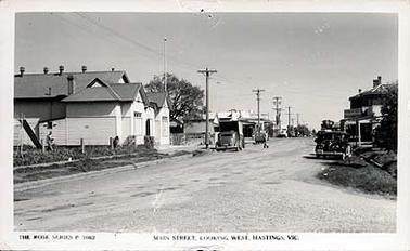 This image shows Main Street, Hastings, looking west. Hastings Hall is in the left foreground and the hotel is on the right. Several cars are parked on either side of the road, there is a man walking beside the hall and another crossing the street. Stevens Bros. store is on the corner of Main Street and Salmon Street.