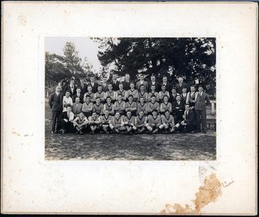 This image shows a football team and its staff, posing for a photograph in front of a large tree. There are twenty men standing and sitting, dressed in suits, white shirts and ties.  One man is dressed all in white.  There are twenty-four footballers standing and sitting in three rows.  There are eight players standing, eight players sitting on a stool and eight players sitting cross-legged on the grass.  All players have their arms crossed except for one.  There is a football on the ground between two players which reads: “Premiers 1928 M.P.F.A.”.  A post and rail fence can be seen behind the men as well as other trees in the background.  The photographers details are printed on the mounting on the right-hand corner.  A crown and the writing ‘by special appointment’ is embossed on the top left-hand corner of the mounting.  