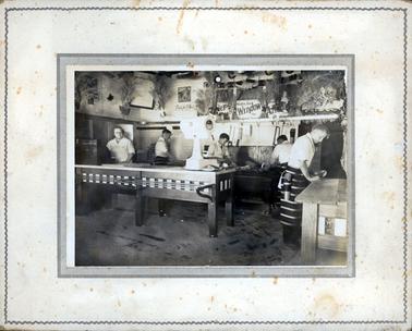 This image shows a butcher shop with four men working on bench tops and a woman standing at the cash register.  The men are wearing white shirts and stripped aprons with a knife belt and holster around their hip.  A large white set of weighing scales sit on the end of the front counter.  The walls are decorated with bunches of dried plants, signs and pictures.  Sawdust is on the floor.  