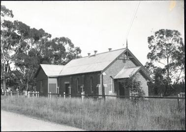 This is an image of a brick church with a galvanised iron roof.  A small building is joined to the back of the church.  A post and rail fence is shown along the right-hand side of the church with a gate half way along.  There is long grass in the foreground and large trees to the left and right of the church.  A small section of road is visible.  