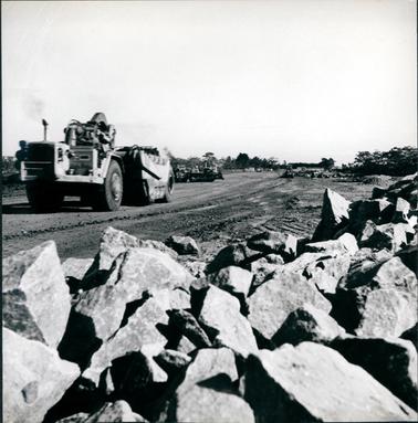 This image shows work on a construction site.  A man is driving a large excavator in the foreground and a bull dozer, grader and several machines can be seen in the background.  Very large rocks are in the foreground.  Trees are seen surrounding the site in the distance.