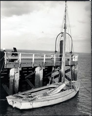 This image is of a small wooden boat beside a pier.  It has a tall mast with a sail lying in the boat.  There are several ropes in the boat.  People can be seen on the left-hand side of the pier.  The pier has white railings and four sets of posts can be seen beside the boat. 