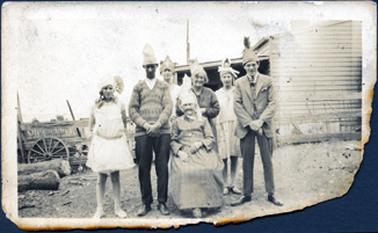 This image shows a group of seven people posing for a photograph.  An elderly woman is sitting on a chair.  She is wearing a long sleeved dress to the ground.  A woman, two men and three young people are standing behind her.  They are all wearing party hats.  The man on the left is wearing a patterned pullover and the man on the right is wearing a three-piece suit.  Two of the girls are wearing knee length dresses.  Part of a house and a tank can be seen behind the group.  A wagon wheel is leaning against a post and rail fence on the left-hand side of the image.  Two large logs are lying on the ground in front of the wheel.  