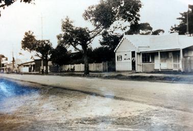 This image shows a line of houses and shops with a street in the foreground running across the width of the photograph.  The weatherboard building on the right-hand side has a three post verandah at the front with a large sign board across the top.  The peaked-roofed section on the left-hand side has a scalloped trim, windows and a door across the front and windows at the side.  A long paling fence is on the left-hand side of the building and shops can be seen further along the street.  Two large trees are on the footpath.
