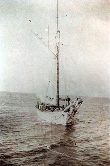 This image shows a wooden sailing boat in the middle of the sea.  It has one very tall mast.  Several people can be seen on the boat.  Part of a smaller boat can be seen in the background.  