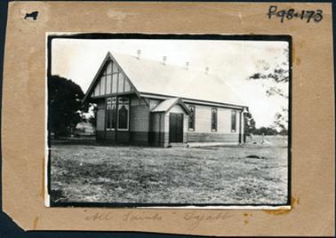 This image shows a church with a large section of grass in the foreground.  A large tree is to the right of the church and other trees can be seen in the background.  The building is timber with an iron roof.  There is a band of darker boards around the bottom of the building. Three windows can be seen on the right-hand side of the narthex and three windows can be seen at the front of the building.  Thin strips of timber  decorate the triangular roof line above the windows.  Four air vents are along the top of the roof.  Part of a post and wire fence can be seen behind the church on the right-hand side.  Written on the bottom of the mounting is: “All Saints Tyabb”.  The mounting is brown paper.