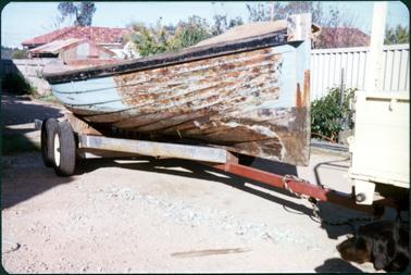 This image shows a ‘rusty’ looking small boat on a trailer.  It is parked on a gravel strip beside a white fence which has a garden in front of it.  Roofs of houses are seen above the fence.  