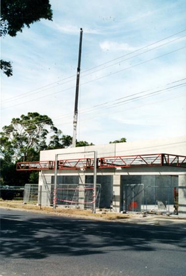 This image shows a construction side and a partly built concrete building.  The top of a tall crane can be seen behind the building and a bitumen street is in the foreground.  Two sets of power lines are across the top of the building and trees can be seen on the left-hand side of the image.  A large shadow from a tree is across the street.  Windswept clouds are in the top half of the image.  