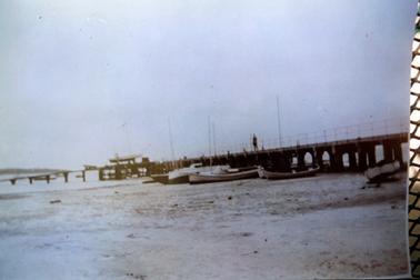 This image shows a jetty reaching out into the water. Several boats are moored at the left of the jetty.  The tide appears to be low as the boats are resting on the sand.  One man is walking along the jetty.  Note the working slipway.  