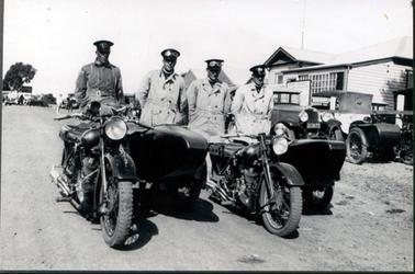 This image shows four men dressed in motorbike outfits standing, with hands behind their backs, behind two motorbikes with sidecars.  They are all wearing flat top hats, goggles and overcoats with belts.  Part of two old model motor cars can be seen at the right-hand side of the image, parked in front of a timber building.  The number plate on one of the cars reads ‘163671”.  There is a large signboard in front of the building.  Other vehicles and people can be seen in the distance.  A large tree is on the far left-hand side in the distance.  