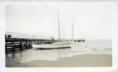This image shows a jetty with several fishing boats moored on the right-hand side of the photograph.  The boats all have tall masts.  The jetty has large dark posts and the railing on the jetty is painted white.  Other masts can be seen in the distance on the other side of the jetty.  The tide appears to be low and there is sand in the foreground.  