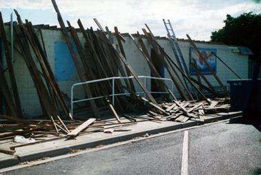 This image shows a cream coloured brick wall with an aluminium ladder and many wooden planks and boards leaning against it and scattered across the footpath. The roof has been taken off.  A painted mural is on the wall and a metal bike rack is against the wall, underneath the mural.  A white pipe railing is in front of the wall.  Part of a large blue bin (skip), is on the right-hand side of the photograph with “No 15” in white printing on the side.  A concrete footpath and gutter and a bitumen street are in the foreground.  Trees can be seen on the far right-hand side of the photograph.