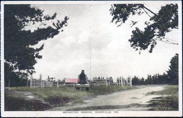 This image shows a large area of grassed land with a fenced area in the centre.  A building with a red roof can be seen in the background and a mounted cannon is in the middle, foreground.  A gravel road is seen on the right-hand side of the image.  Pine tree branches can be seen in the foreground, left-hand side and gum tree branches on the right-hand side of the image.  Pine trees can be seen in the background.  Printed at the bottom is: “Recreation Reserve, Somerville, Vic.”.
