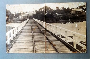 This photograph was taken from a middle of a wooden jetty looking back to the shore.  The jetty has rail tracks down the centre and a white two railing fence on either side.  Water and rocks can be seen on either side of the jetty.  Several buildings and trees are along the shoreline.  A vertical line runs through the centre of the image from top to bottom, the result of a crease in the original photograph.